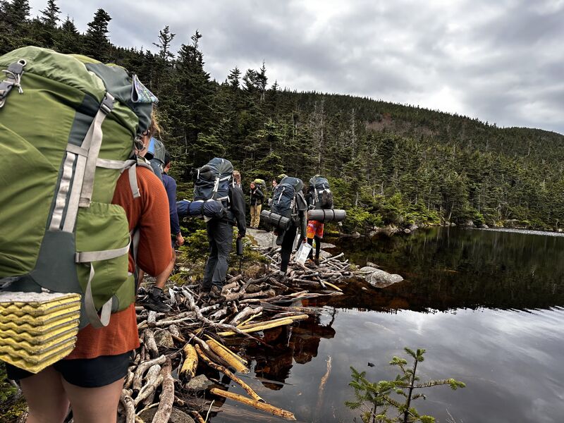 A group of hikers with large backpacks are walking along a narrow, rocky path next to a lake. The path is covered with fallen branches and debris. The lake is calm and reflects the surrounding trees and sky. The sky is cloudy and overcast. The hikers are wearing a variety of clothing, including hiking boots, pants, and jackets. The backpacks are various colors and sizes.